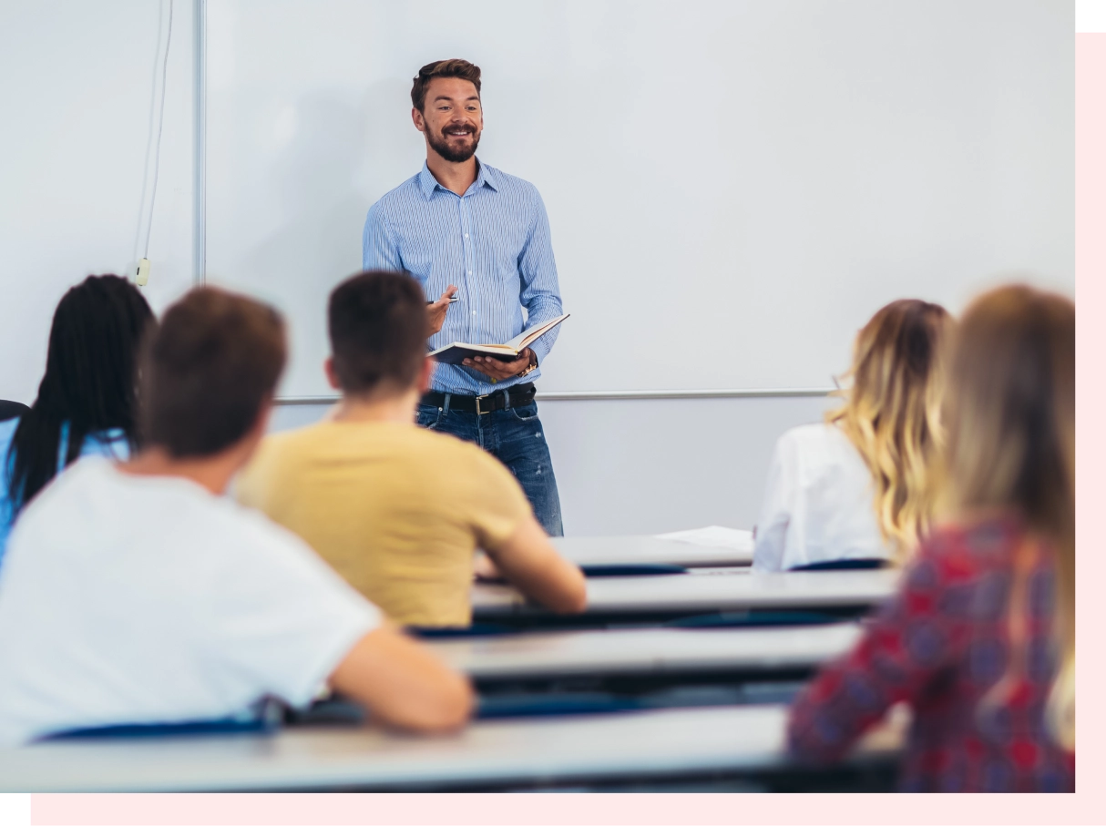 A man stands in front of a classroom, holding a book and speaking to a group of seated students.
