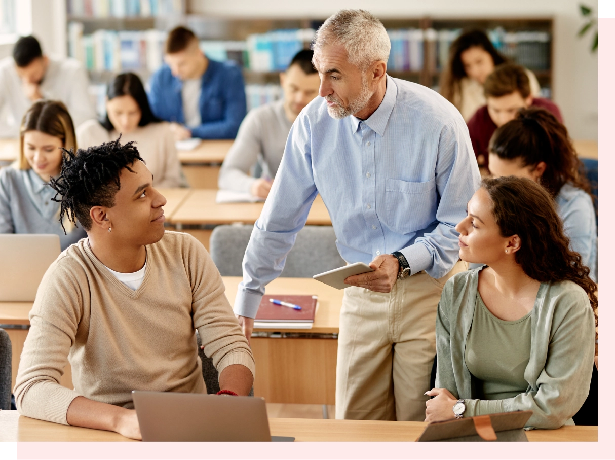 A teacher engages with students in a classroom, using a tablet. Students sit at desks with laptops, appearing attentive. Bookshelves fill the background.