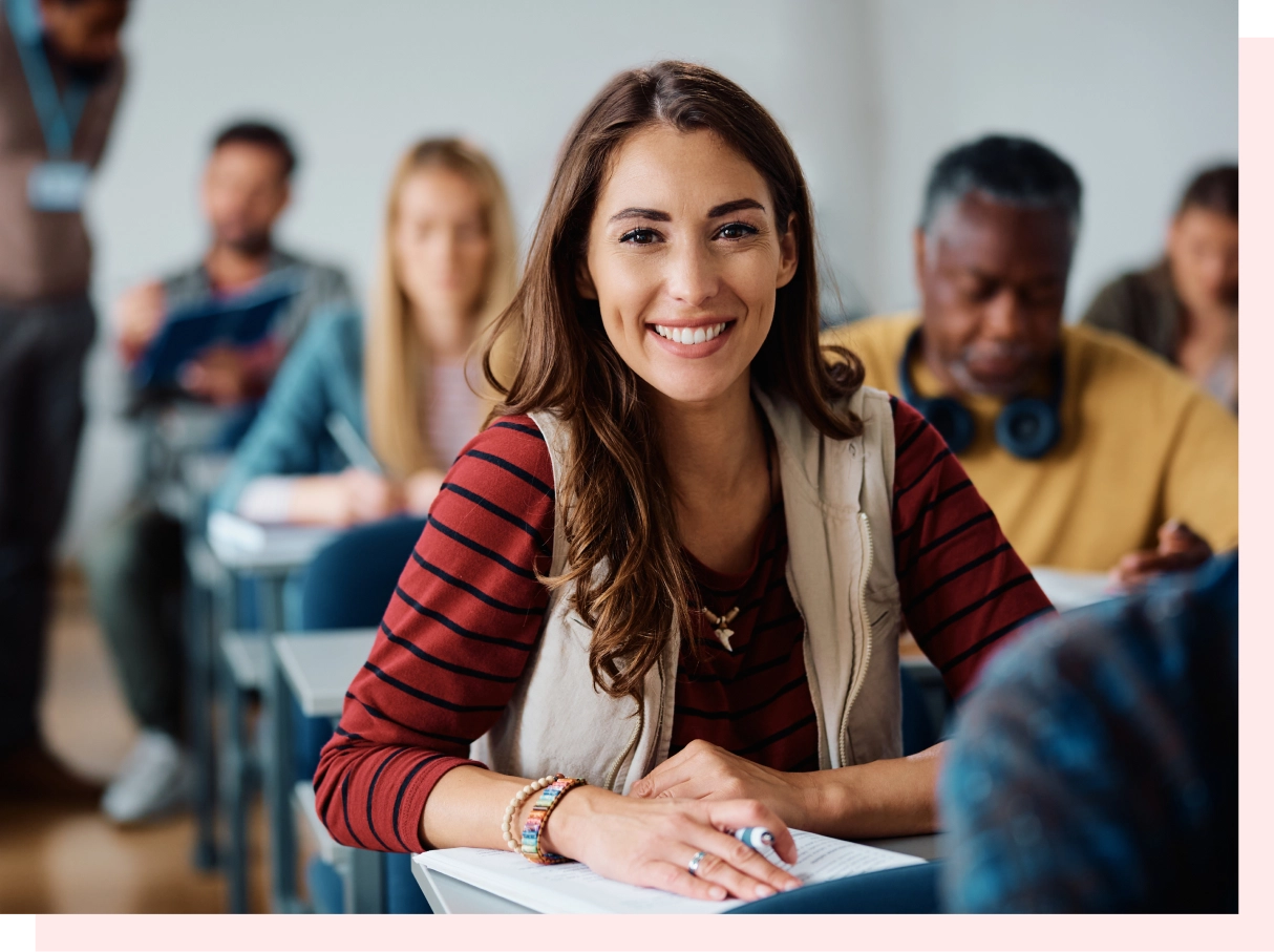 A woman in a classroom smiles at the camera while sitting at a desk. Other students are in the background.
