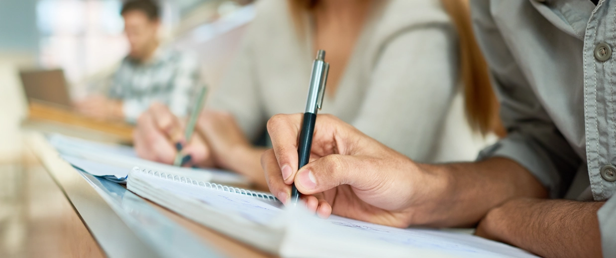 Close-up of two people writing notes with pens in a notebook, with another person blurred in the background.