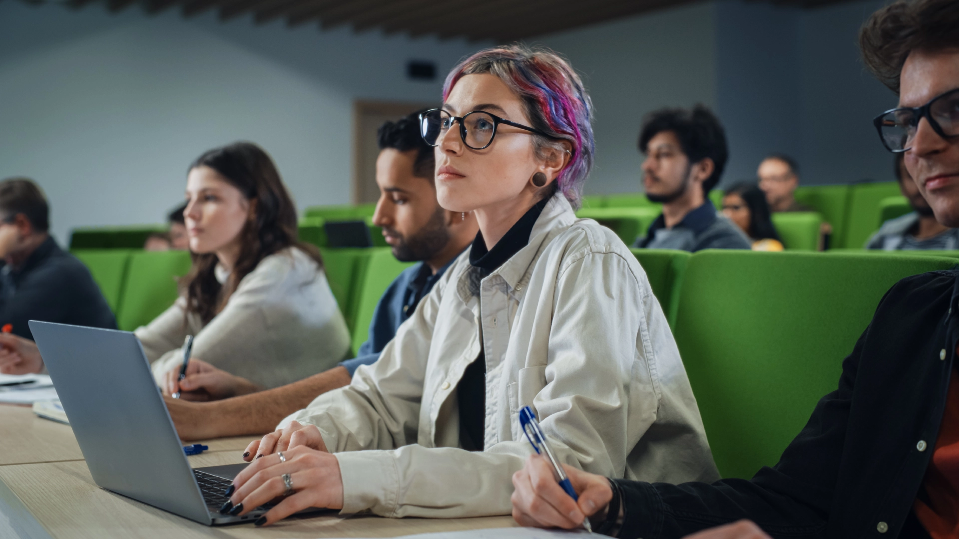 People sitting in a classroom, listening intently and taking notes. One person with colorful hair is using a laptop.