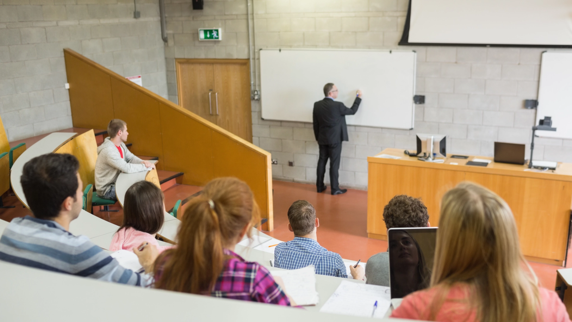 Professor teaching in a lecture hall with students seated in tiered rows, some taking notes and others using laptops.