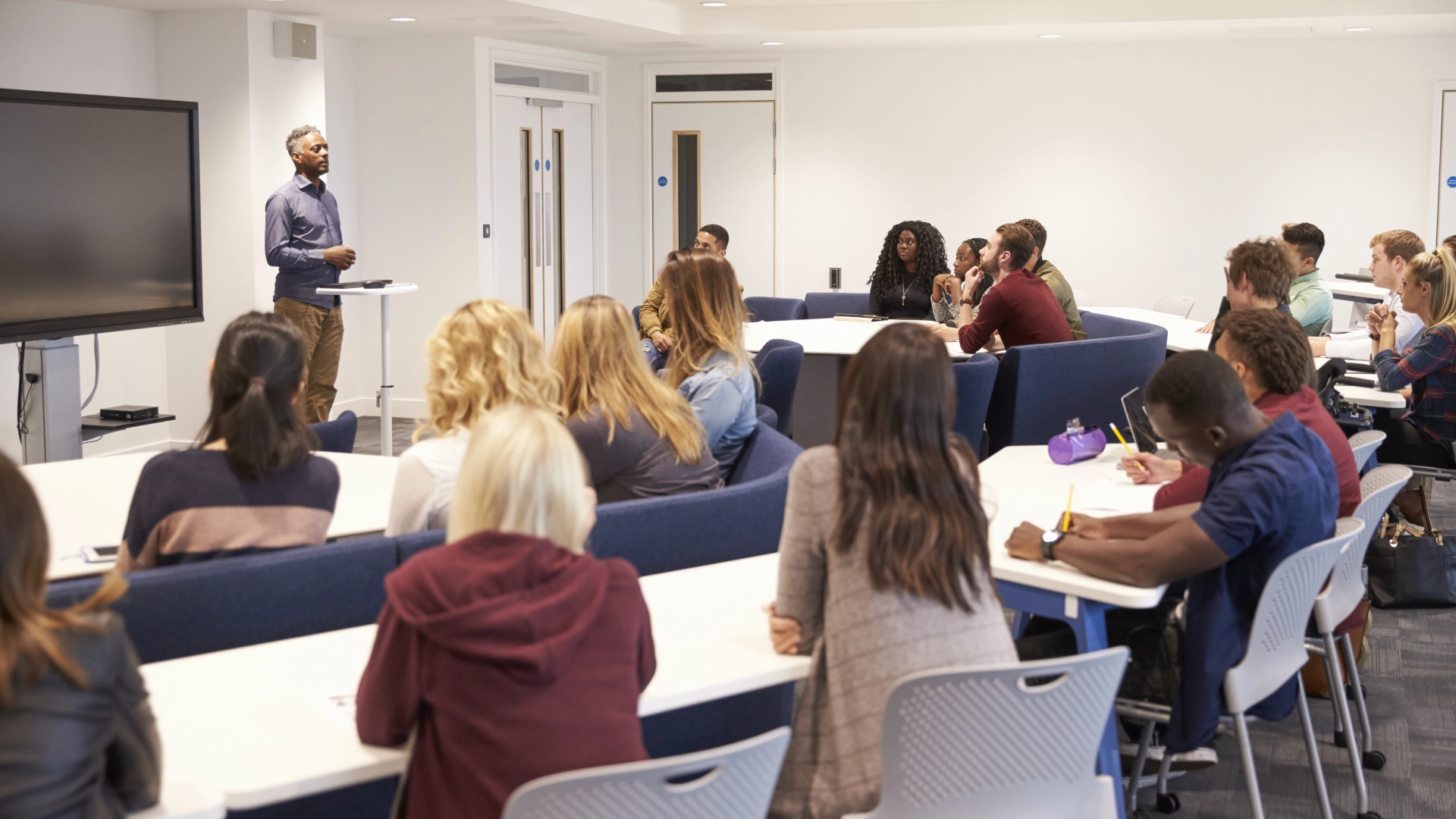 A classroom with a lecturer speaking to students seated at long tables, facing the speaker and a large screen.
